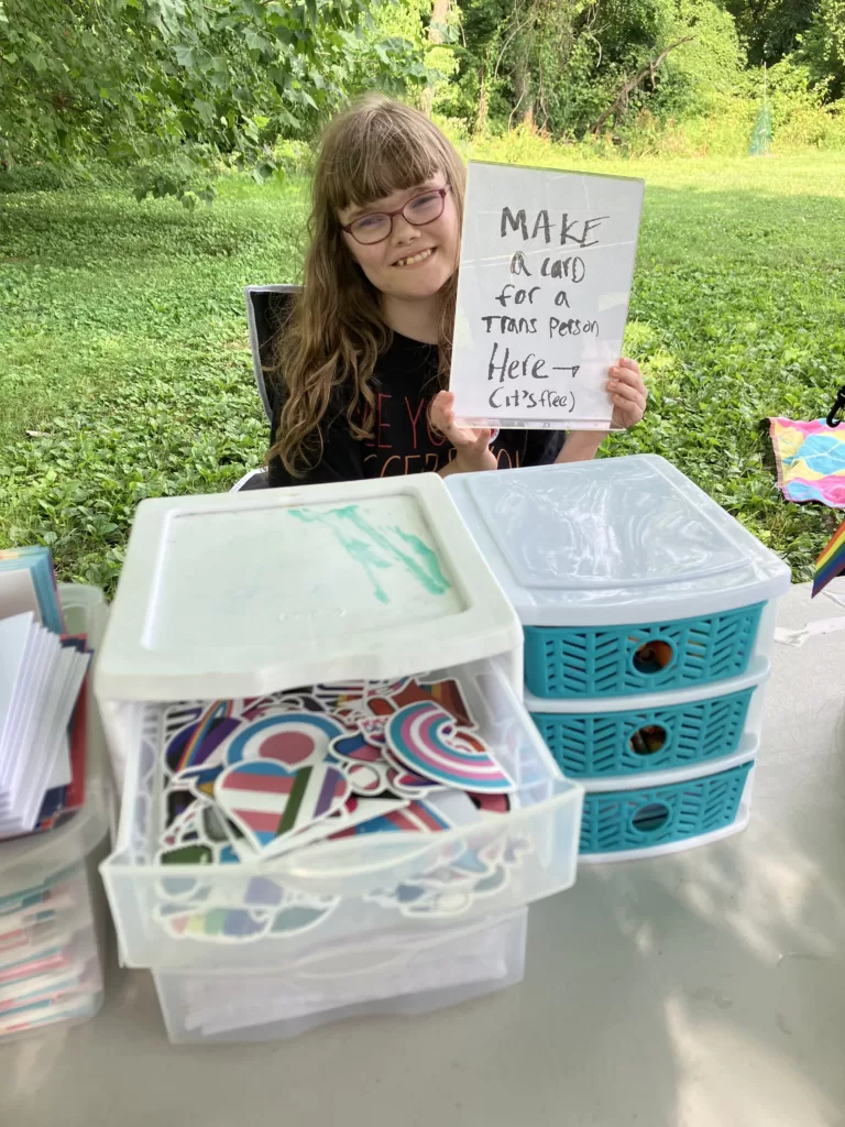 A photo of an LTAP organizer holding a sign that says "Make a card for a trans person here, it's free" while sitting behind the card-making table.